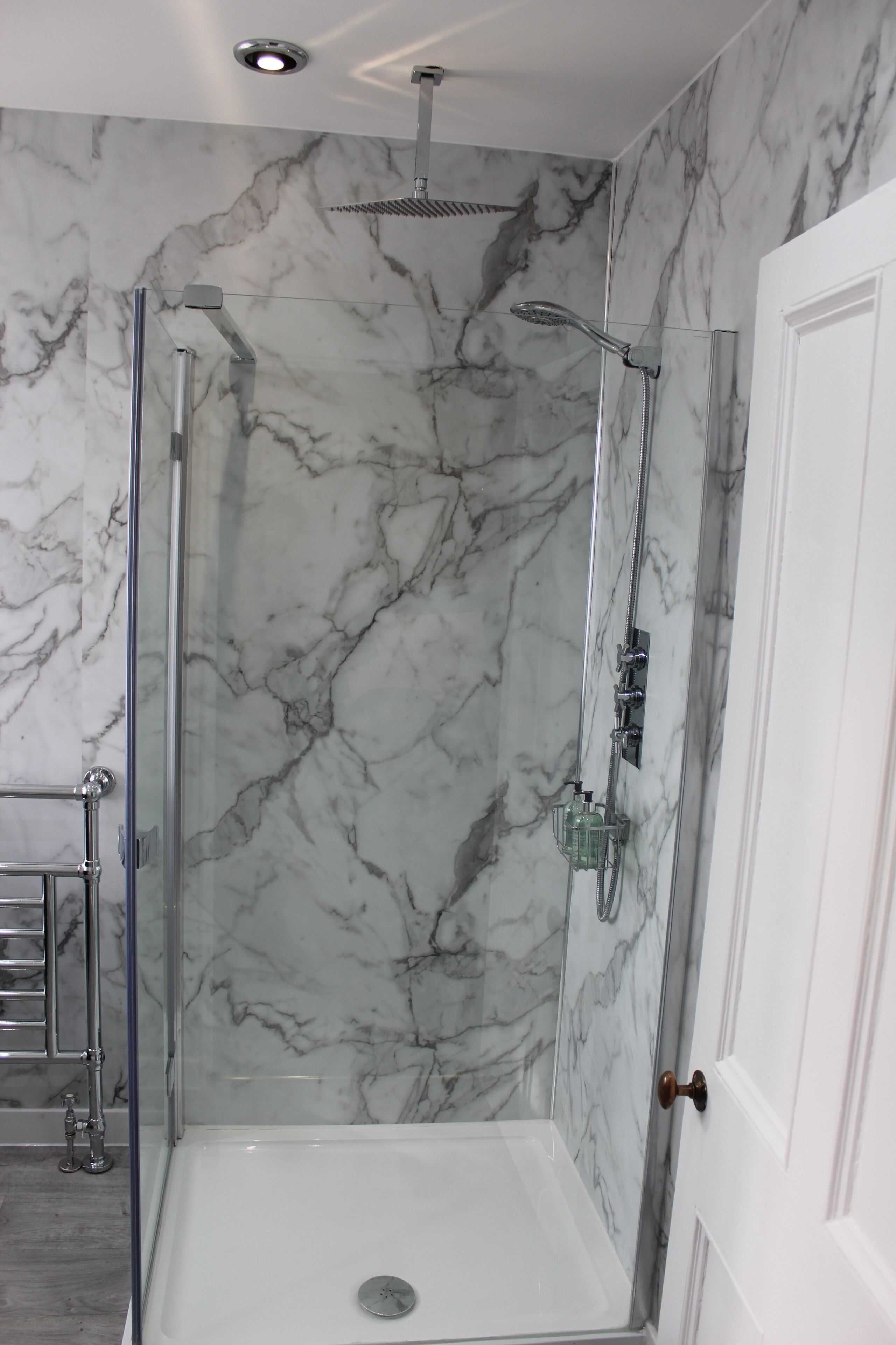 A glass-enclosed shower with a marble wall backdrop, a white door, and a visible showerhead.