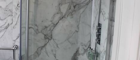 A glass-enclosed shower with a marble wall backdrop, a white door, and a visible showerhead.