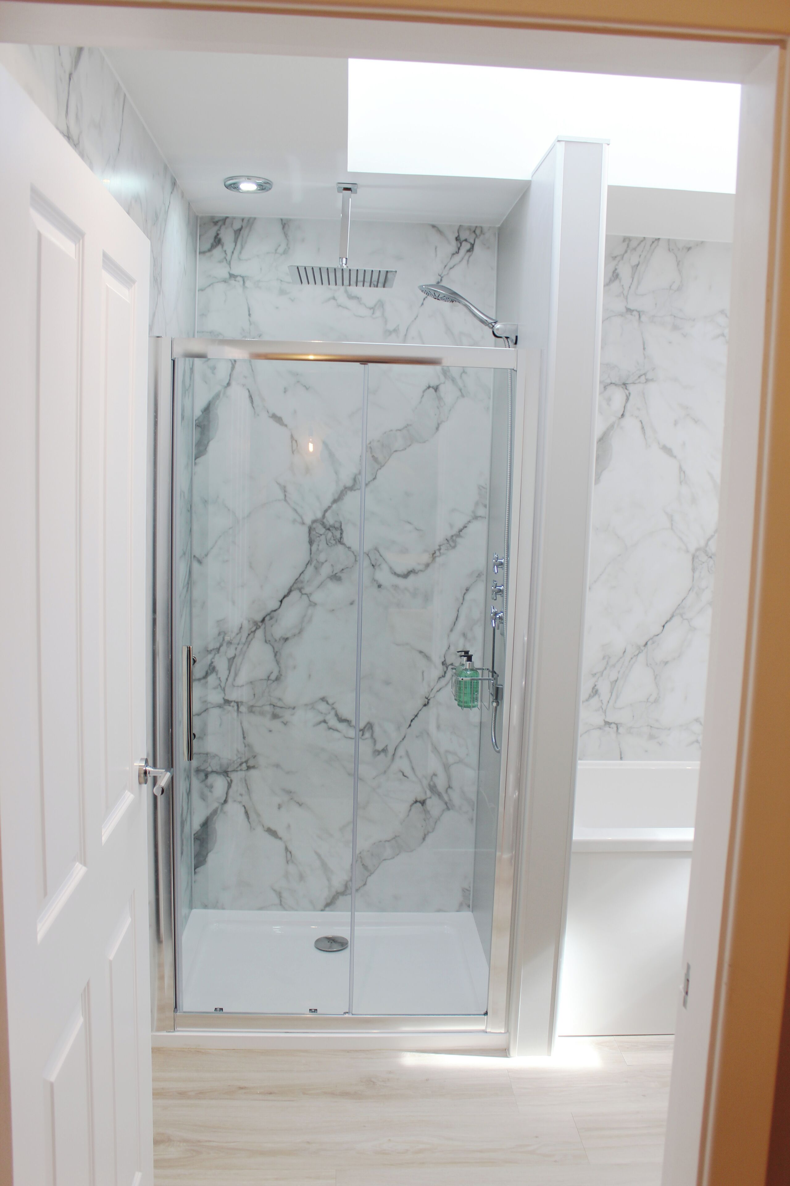 A glass-enclosed shower with a marble wall, a visible soap dispenser, and a ceiling light.