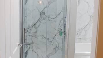 A glass-enclosed shower with a marble wall, a visible soap dispenser, and a ceiling light.