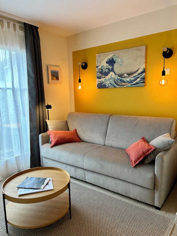 A living room with a grey sofa, two red cushions, a round wooden coffee table, a woven rug, a wall-mounted light, and a Japanese wave painting. - House very close to the beach and coastal path (Locmaria)