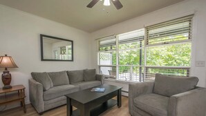 Ein Wohnzimmer mit grauem Sofa, Couchtisch und Lampe. Ein Fenster mit Jalousien und ein Deckenventilator sind ebenfalls vorhanden.