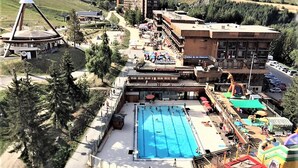 Una piscina con plataforma de salto, un edificio grande de varios pisos y un área de juegos con estructuras inflables.