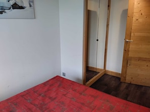 Une pièce avec une couvre-lit à carreaux rouges, une porte en bois et un mur blanc orné d’un tableau encadré.