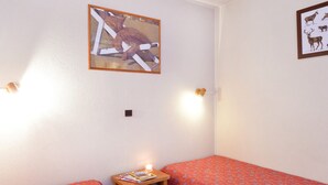 Une petite pièce bien éclairée, avec deux lits simples, une petite table sur laquelle est posée une bougie, et deux tableaux encadrés accrochés aux murs.