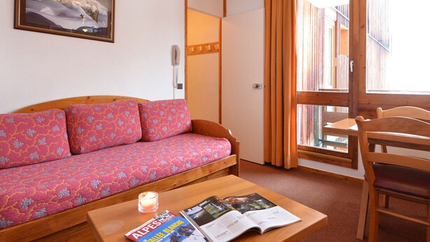 Une pièce chaleureuse avec un canapé en bois, une petite table sur laquelle sont posées une bougie et des magazines, une chaise en bois et une grande fen être avec des rideaux.