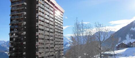 A high-rise building with multiple balconies, surrounded by snow-covered mountains and trees.