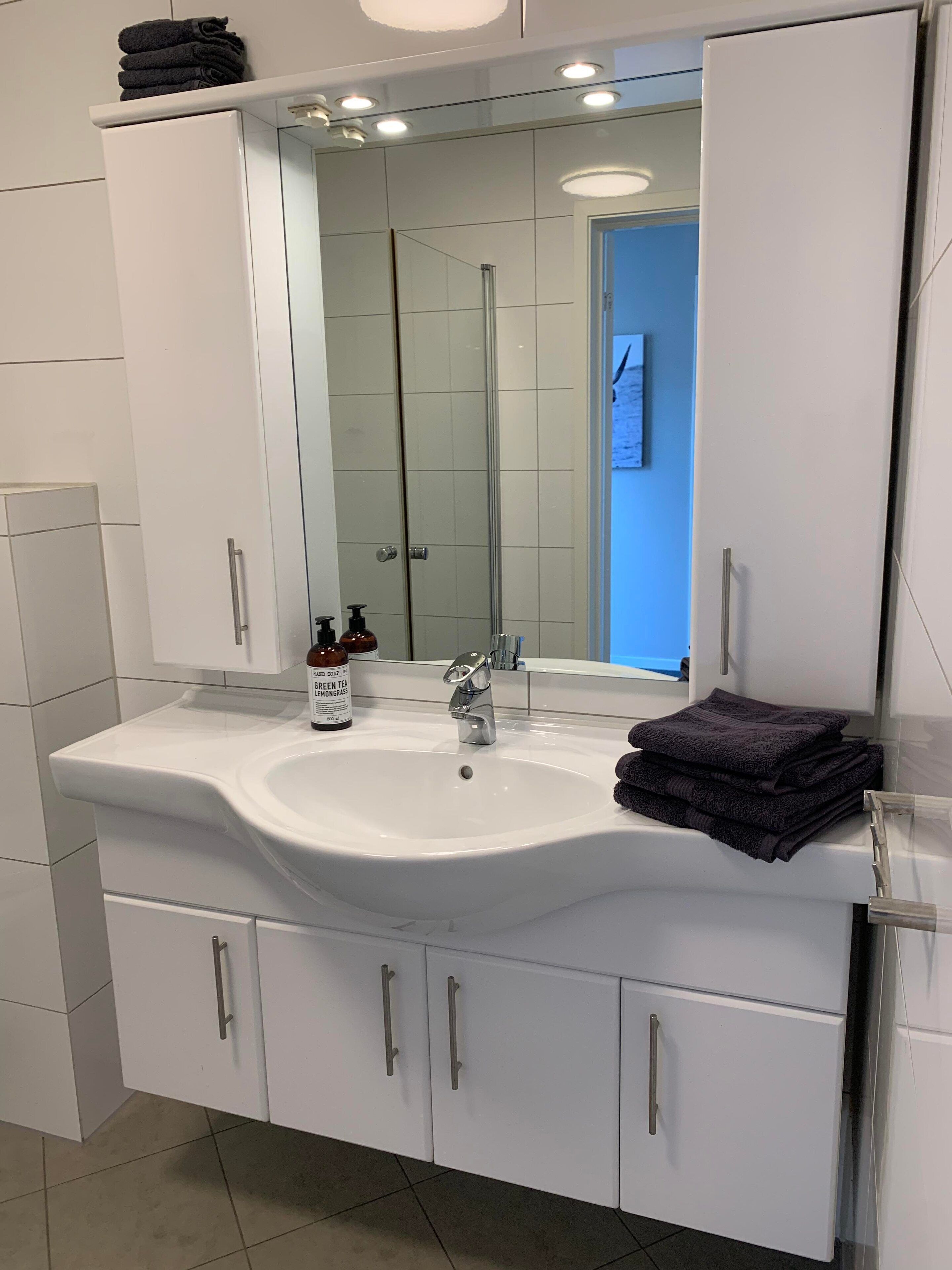 Un baño moderno con un lavabo blanco, un espejo y una ducha a la vista al fondo.
