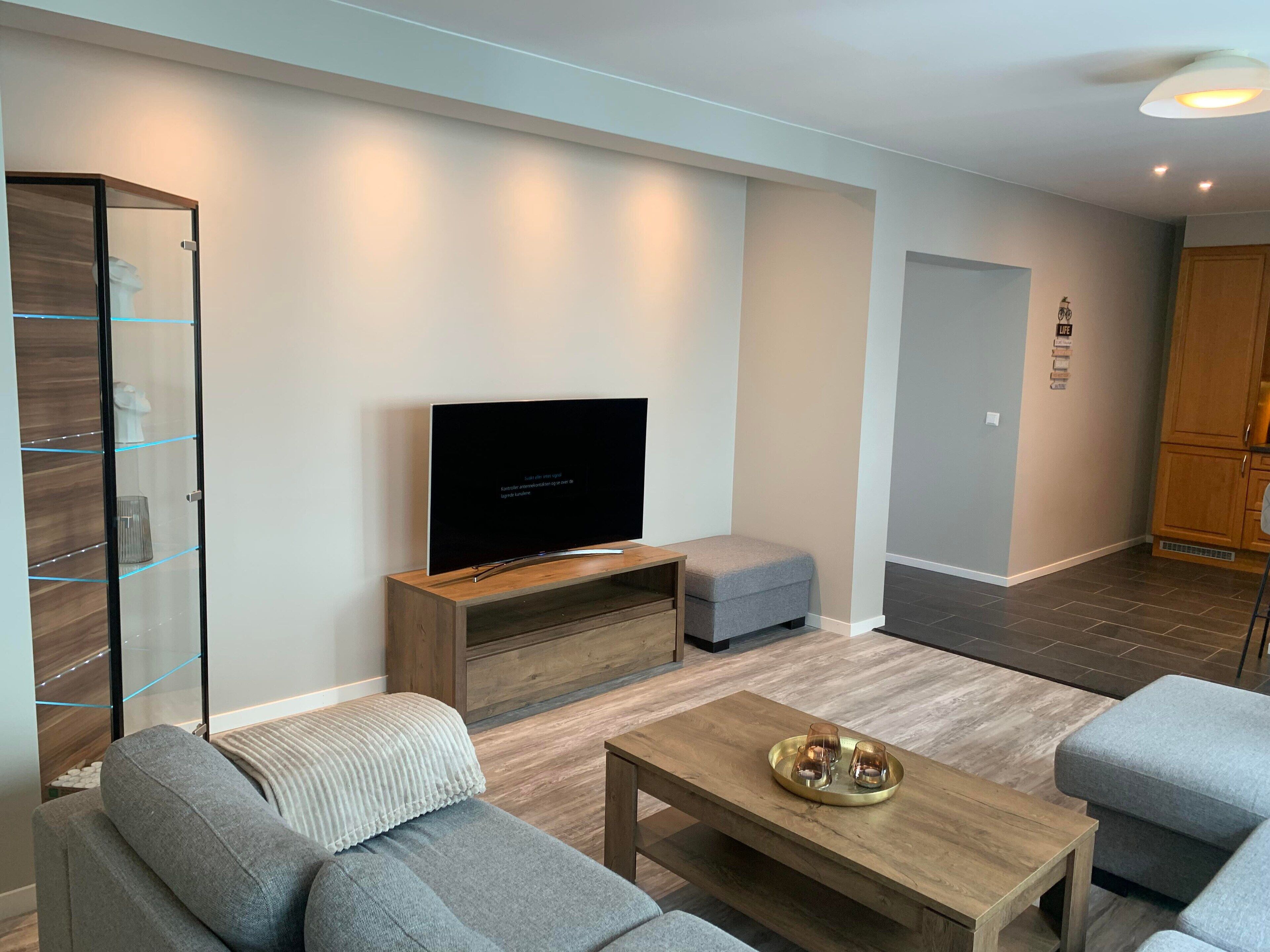 A modern living room with a flat-screen TV, grey sofas, a wooden coffee table, and a glass cabinet.