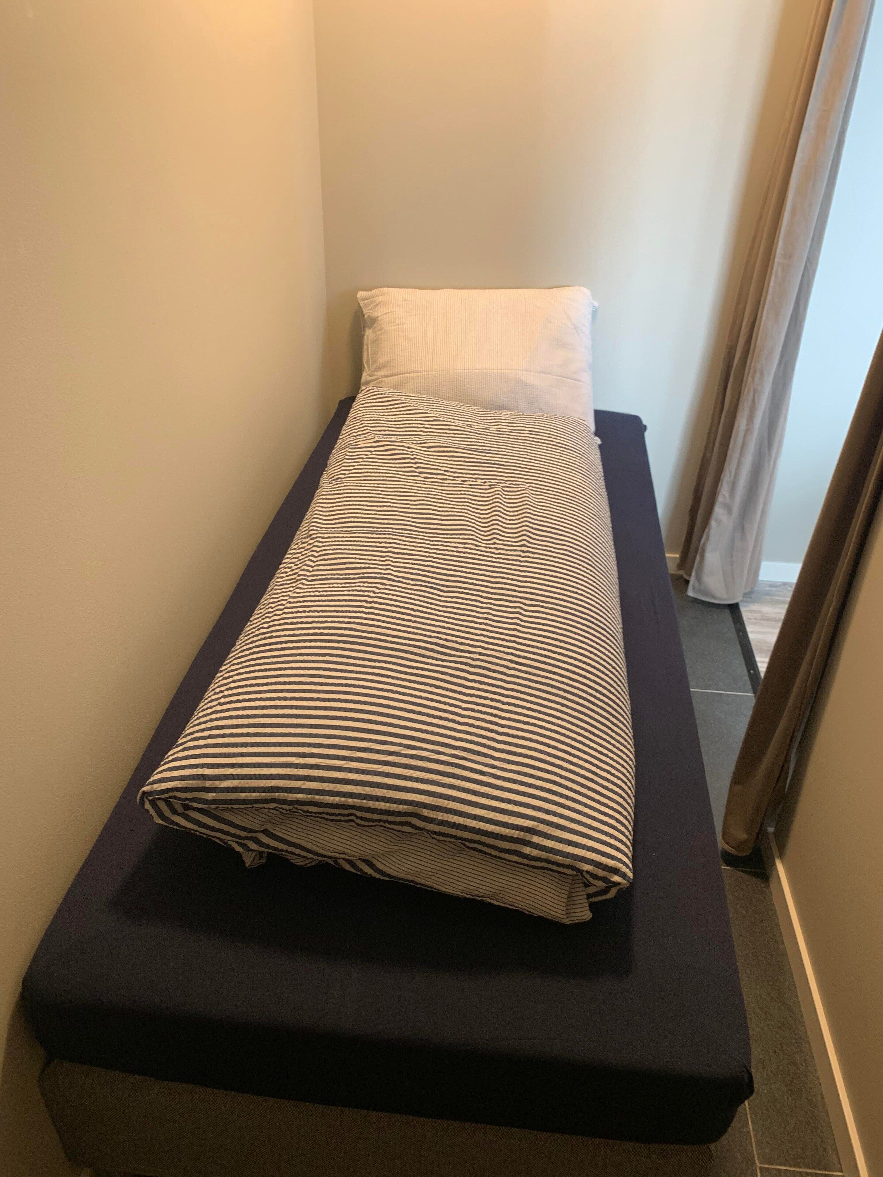 Pieni, kapea huone, jossa on yksi sänky, tyyny ja tumma penkki.