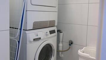 Un espace buanderie compact comprenant une machine à laver et un sèche-linge superposés, un étendoir et une poubelle blanche.