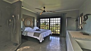 Une chambre à coucher comprenant un lit, un ventilateur de plafond, une grande fenêtre avec vue et une sculpture murale.