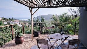 Une terrasse extérieure couverte, avec une table et des chaises, entourée de plantes en pot et offrant une vue sur l’océan.