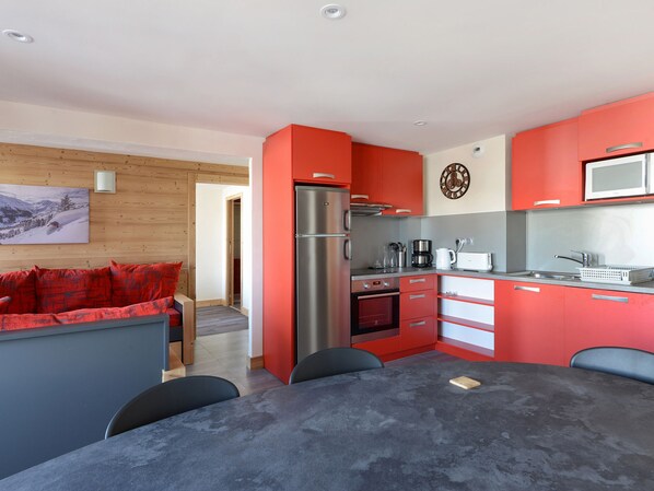 A modern kitchen with red cabinetry, a stainless steel refrigerator, and a microwave. There is a dining table with chairs, and a wooden wall with a picture.