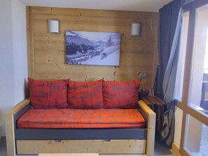 A wooden-framed sofa with red cushions, a wall-mounted picture of a snowy landscape, and a window with curtains. - Renovated studio for 2, ski-in, pets, parking (La Plagne Montchavin Les Coches)