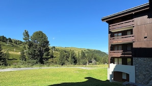 Um edifício moderno com fachada de pedra e varandas de madeira, com colinas verdes e árvores ao fundo.