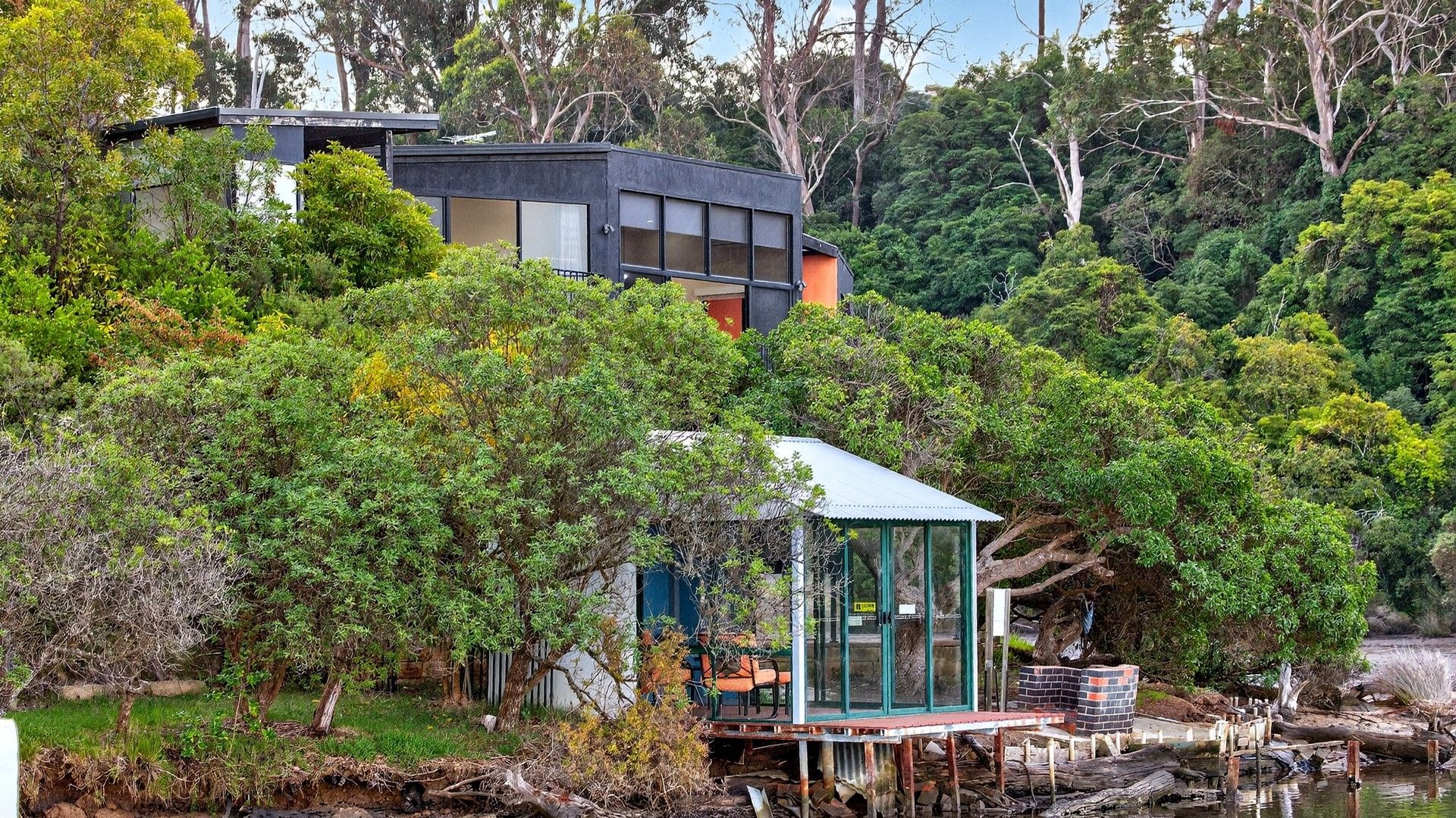 Uma casa moderna com uma estufa de vidro, rodeada por uma floresta densa e um corpo de água.