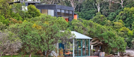 Une maison moderne dotée d’une véranda en verre, entourée d’une forêt dense et bordée par un plan d’eau.