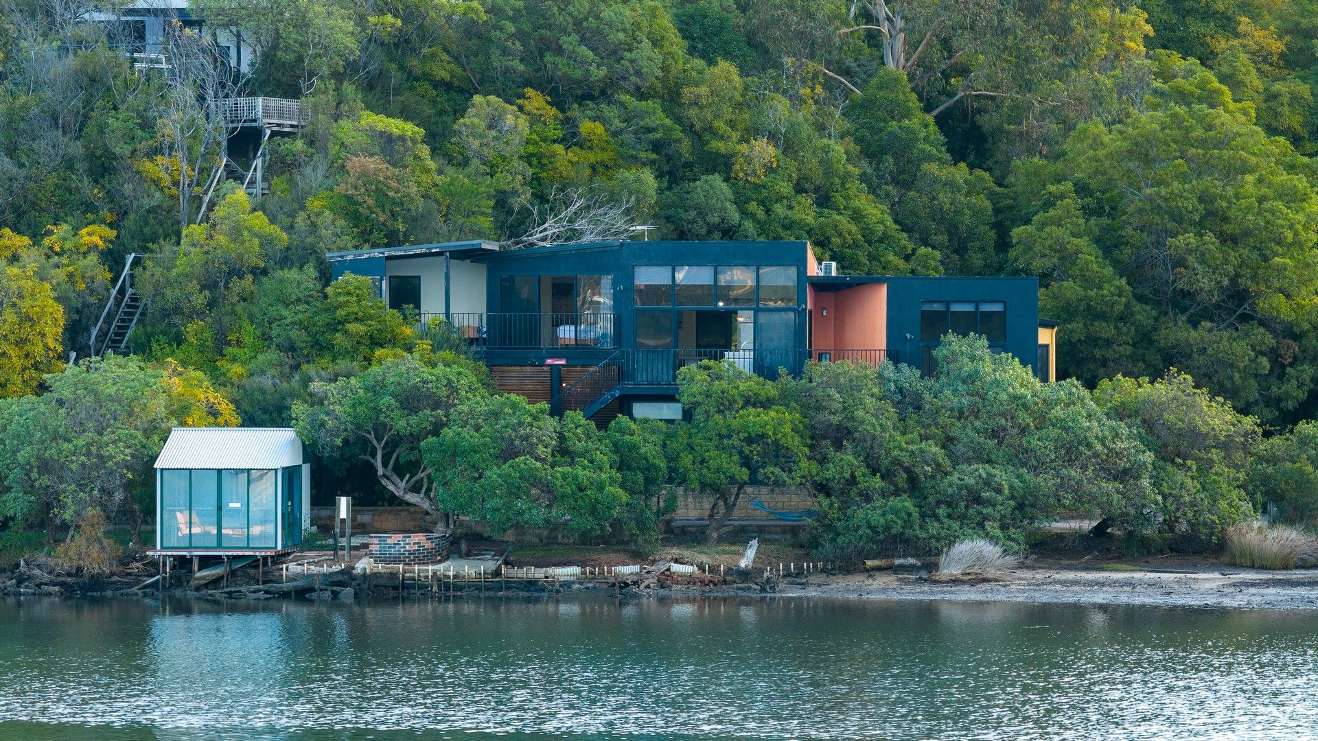 Une maison moderne dotée d’une grande terrasse offrant une vue sur un lac, et entourée d’une forêt dense.