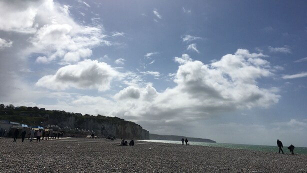 Ein Kiesstrand mit Menschen, die entlanggehen und sitzen, Felsen im Hintergrund und einem klaren Himmel.