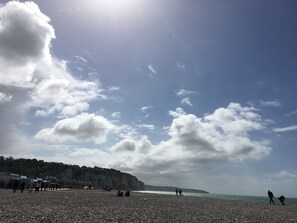 A pebbled beach with people walking and sitting, cliffs in the background, and a clear sky.
