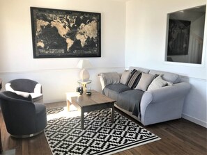 Un salon moderne avec un canapé gris, un tapis à motifs noir et blanc, une table basse ronde, un fauteuil gris et une œuvre d’art représentant une carte du monde accrochée au mur.