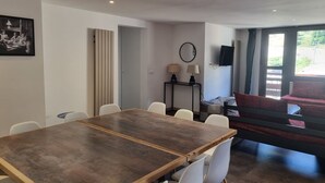 Un espace repas moderne avec une table en bois et des chaises blanches, une petite table d’appoint avec des lampes, un téléviseur fixé au mur et un canapé rouge en arrière-plan.