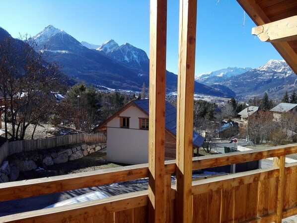 A wooden balcony with a view of a small house and snow-covered mountains.