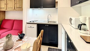 Et lille køkken med vask, komfur og mikroovn. Der er en rød sofa og et spisebord med en vase og skåle.