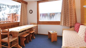 A cozy room with a wooden table and chairs, a small wooden stool, a sofa with patterned cushions, and a window with curtains.