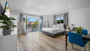 A modern bedroom with a large bed, a dining area with blue chairs, and a balcony with a view of houses. - Studio Beach Rental in Pacific Beach, San Diego (San Diego)