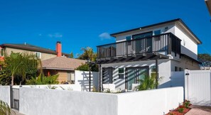 A modern two-story house with a balcony, white exterior walls, and a gated entrance.