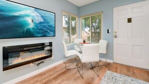 Een moderne woonkamer met een open haard, een eethoek met witte stoelen en een grote tv waarop een surfer op een golf te zien is.