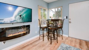 A living room with a fireplace, a TV displaying a surfer, and a dining area with chairs and a table. - Beach & Park Family Fun Getaway -Steps to Sand (San Diego)