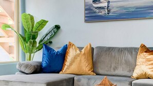 A modern living room with a grey sofa, blue and gold cushions, a potted plant, and a painting of a boat on the wall. - Beach & Park Family Fun Getaway -Steps to Sand (San Diego)