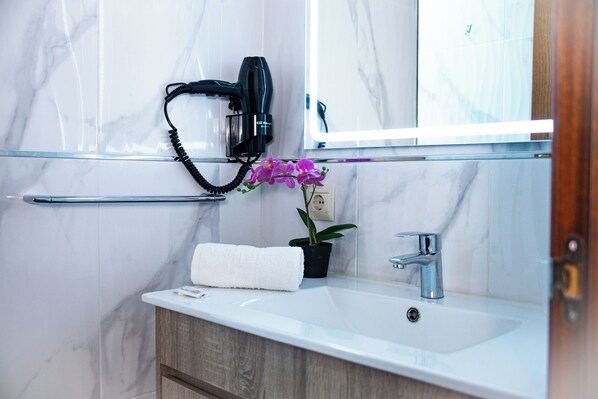 Une salle de bain avec un mur en marbre, un lavabo blanc, une serviette, une orchidée en pot et un sèche-cheveux fixé au mur.