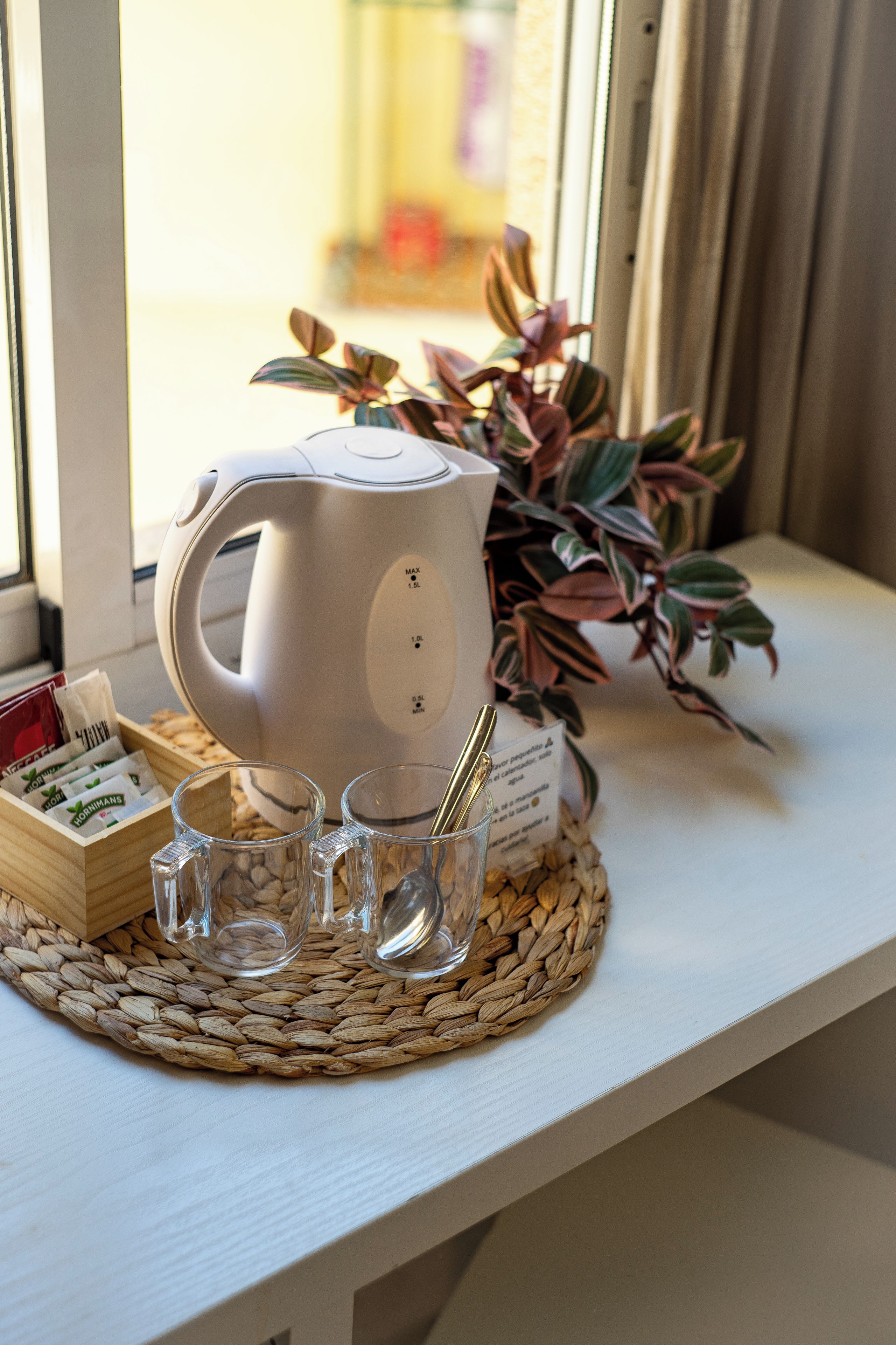 Una tetera eléctrica blanca sobre un posavasos tejido, una taza de vidrio, una cuchara y una caja de bolsitas de té en un alféizar.