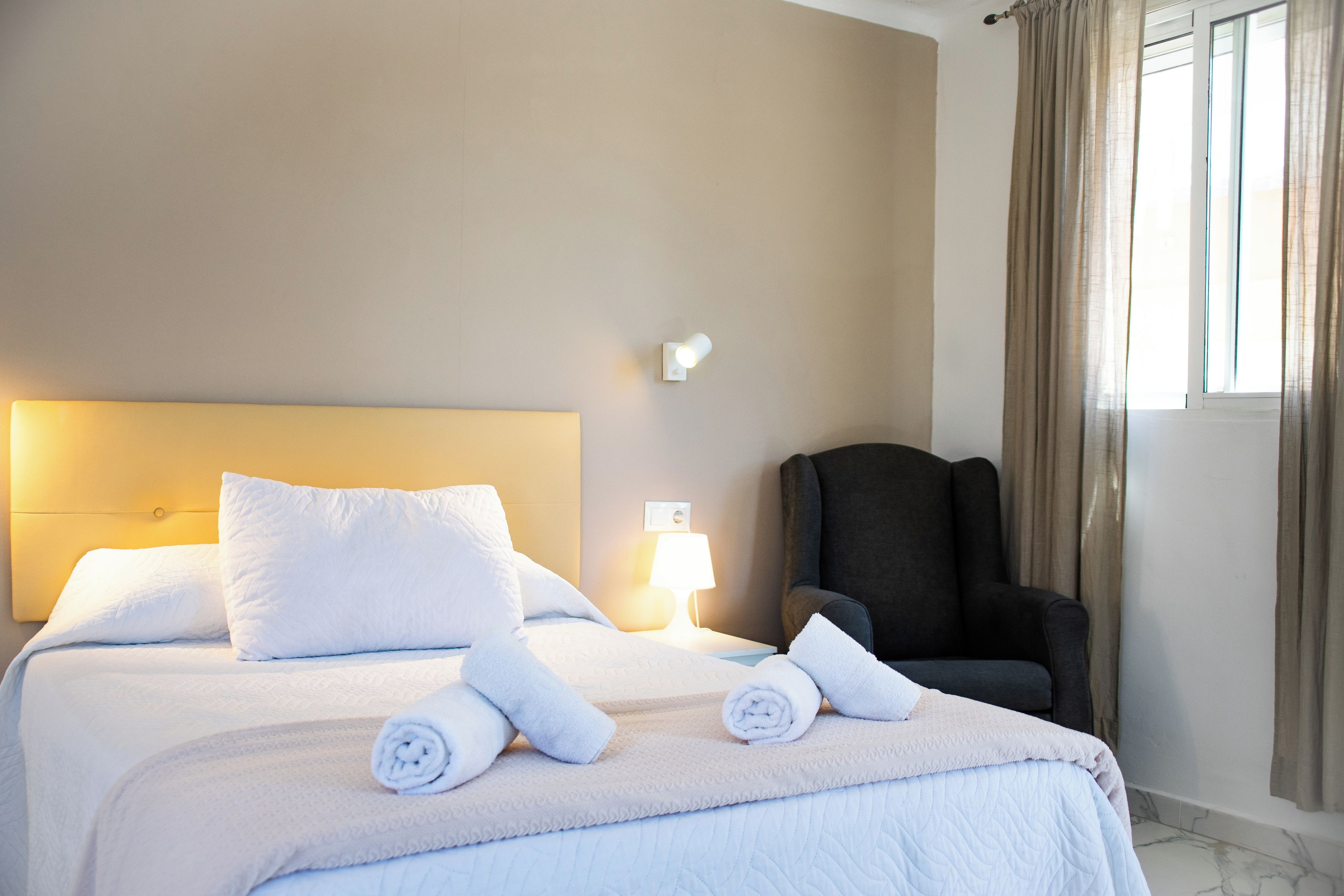 Una habitación de hotel con cama, almohadas, una silla, una lámpara y una ventana con cortinas.