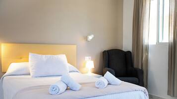 Una habitación de hotel con cama, almohadas, una silla, una lámpara y una ventana con cortinas.