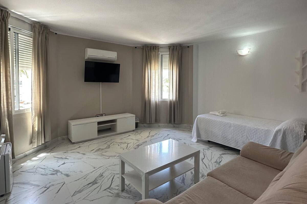 A modern hotel room with a marble floor, a flat-screen TV mounted on the wall, a bed with a white duvet, a beige sofa, and a small white table.