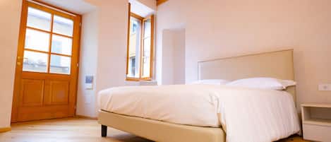 A bedroom with a wooden floor, a bed with white bedding, a nightstand, and a window with a view of buildings.