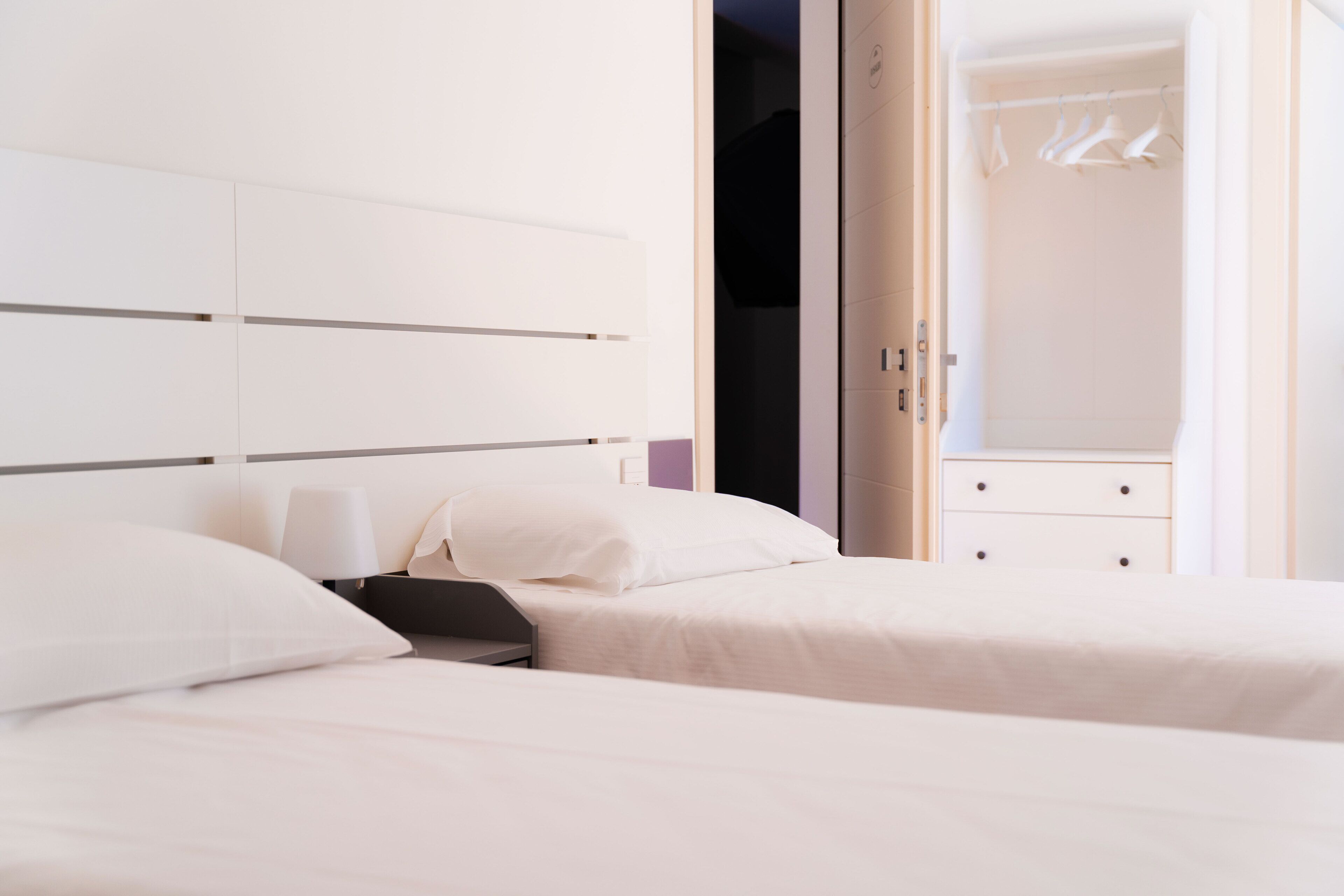 A modern hotel room with two beds, a wardrobe, and a bathroom visible through an open door.