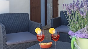 Une terrasse avec des meubles en osier, deux verres d’une boisson rouge agrémentée de tranches d’orange, un bol de chips et un pot de lavande.