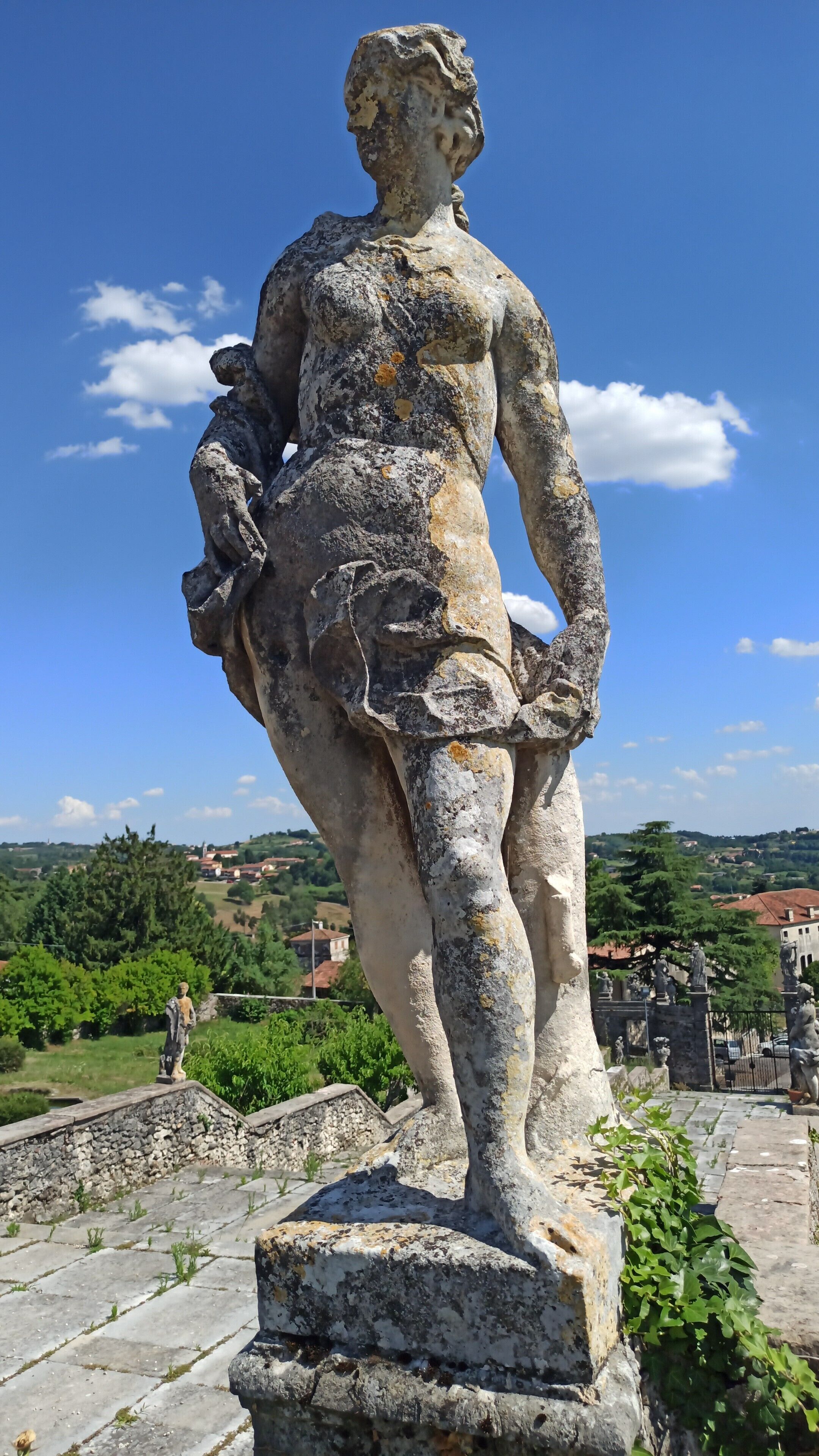 A stone statue of a woman with a child, standing on a pedestal in an outdoor setting with a clear sky and distant buildings.