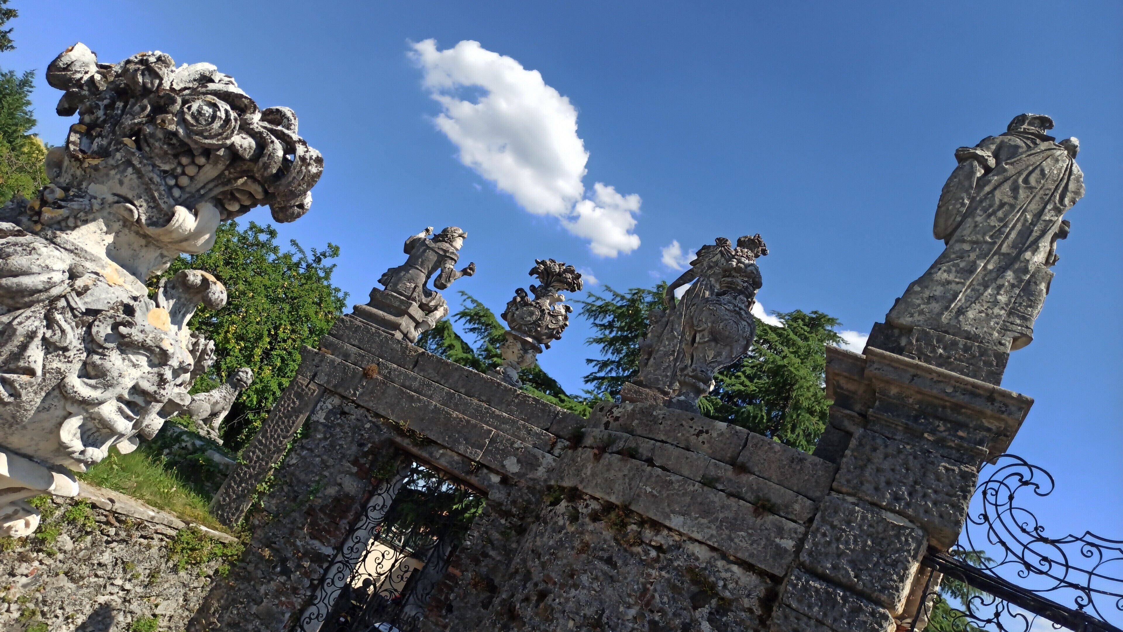 Un arco in pietra con elaborate decorazioni scolpite e statue, che conduce a una rigogliosa area verde.
