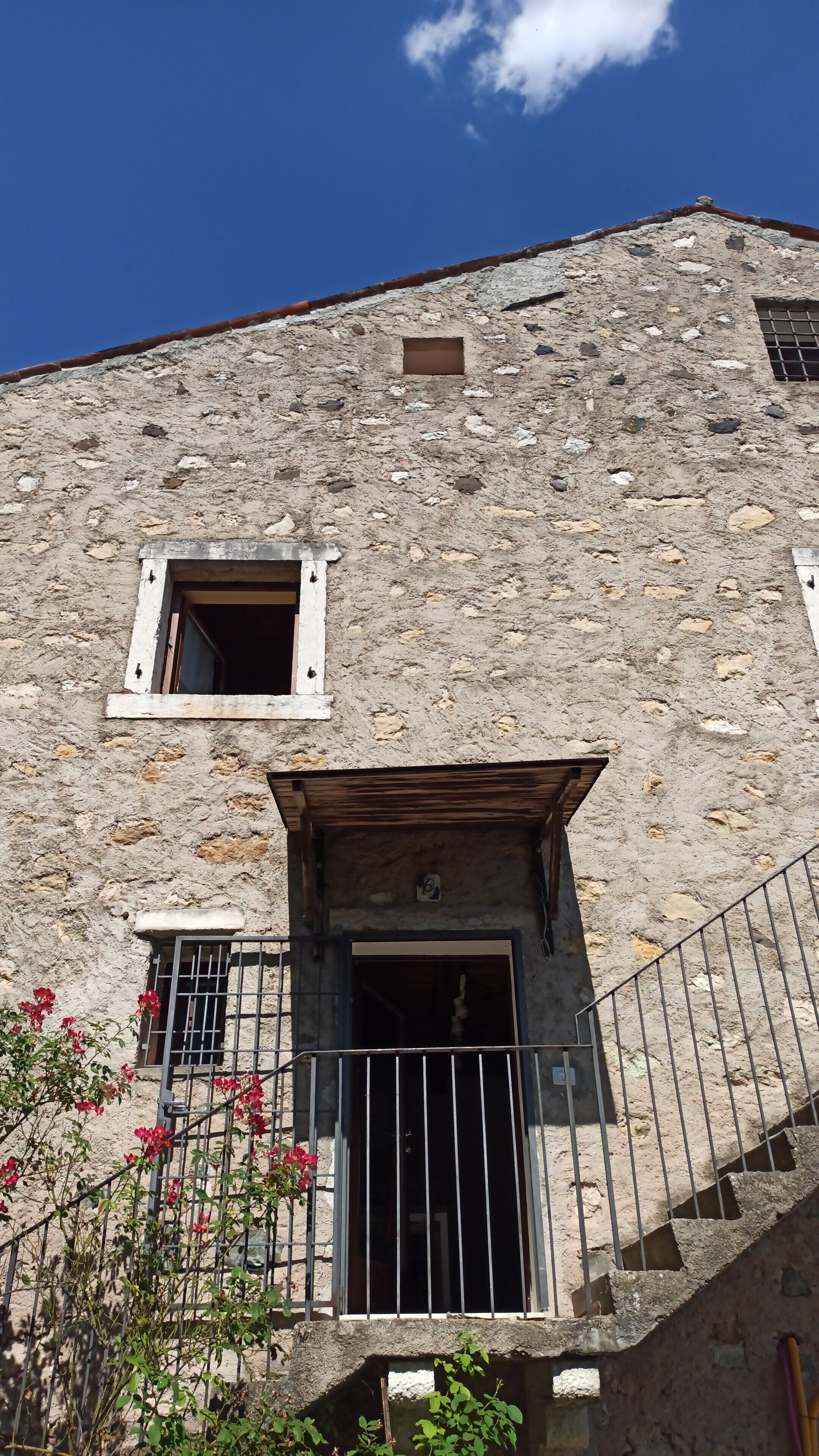 A rustic stone building with a small window and a wooden door, surrounded by a metal railing and some greenery.