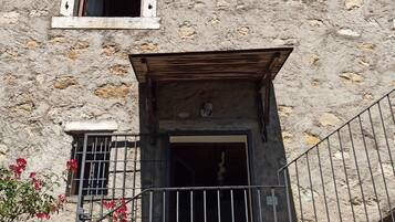 A rustic stone building with a small window and a wooden door, surrounded by a metal railing and some greenery.