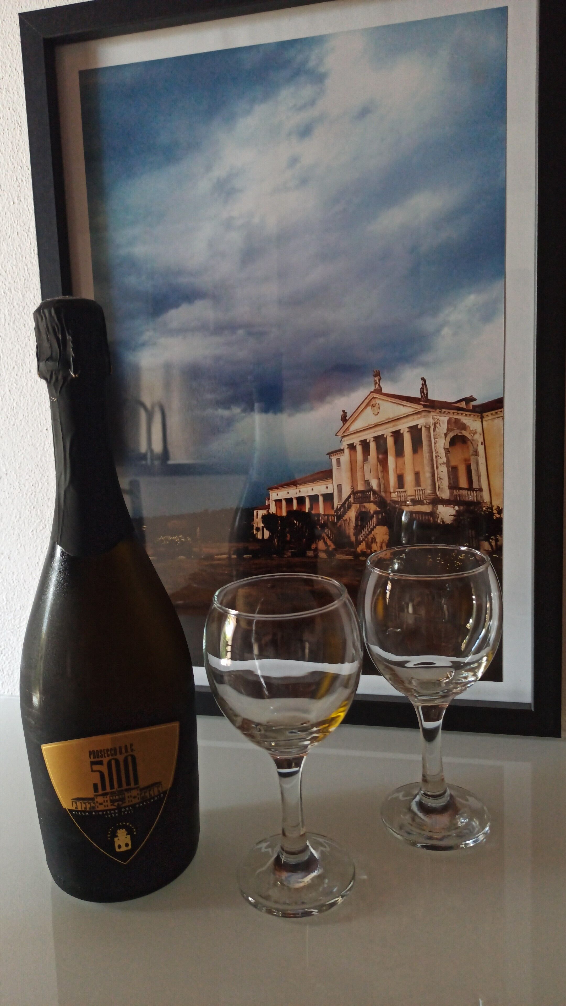 Une bouteille de Prosecco 500, deux verres à vin vides et une photo encadrée représentant un bâtiment avec des colonnes et un escalier.