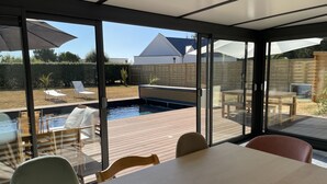 Ein moderner, überdachter Poolbereich mit Glaswänden, Holzterrasse und Sitzgelegenheiten im Freien.