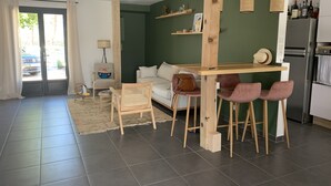 Ein modernes Wohnzimmer mit einer Couch, einem Esstisch mit Stühlen, einer Küchenzeile inklusive Kühlschrank und einem großen Fenster mit Vorhängen.